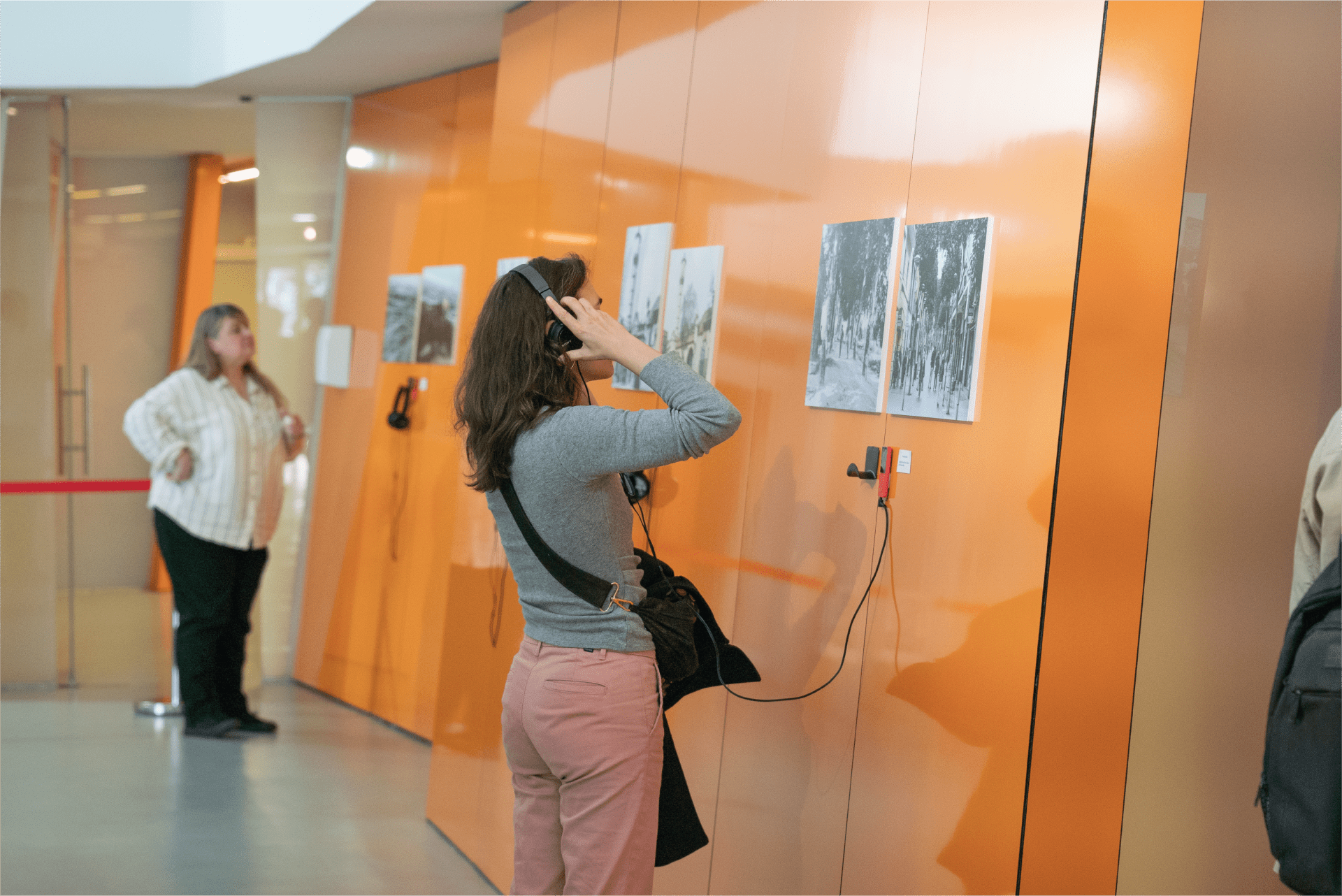 Una chica mira dos fotos colgadas en la pared y escucha un audio con unos auriculares que cuelgan directamente de la pared.