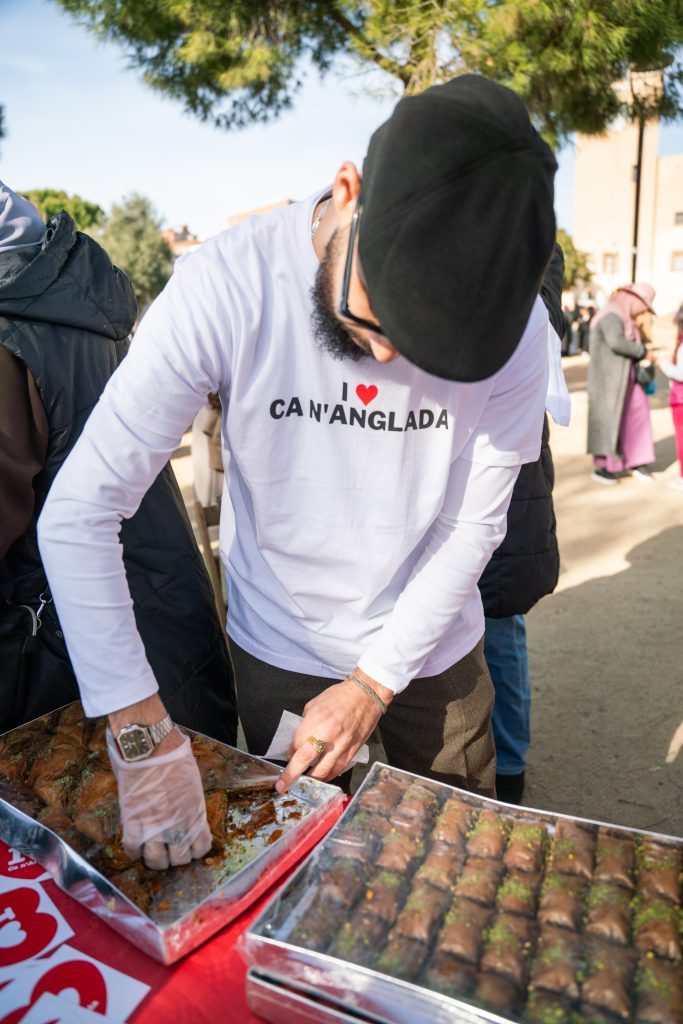 Un noi amb boina i una samarreta on hi diu "I Love Ca n'Anglada" reparteix pastes àrebs en una paradeta de menjar situada en una plaça.